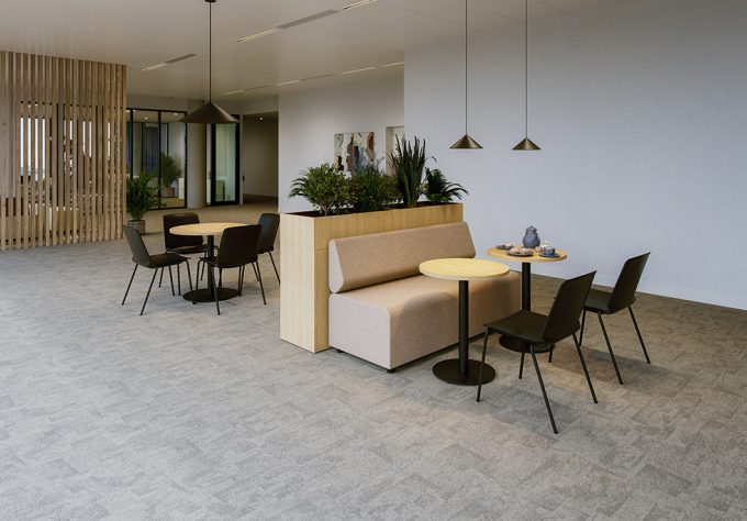Modern lounge area with light-colored carpet flooring and an open layout. In the foreground, there is a beige upholstered bench positioned in front of a low plant divider in light wood finish. Several green plants grow from the divider, visually structuring the seating area. In front of the bench are two round tables with light tabletops and black bases, surrounded by matching black chairs. Simple black pendant lights hang above the seating areas. In the background, another table-and-chair grouping and a partition made of vertical wooden slats are visible, helping to structure the space.