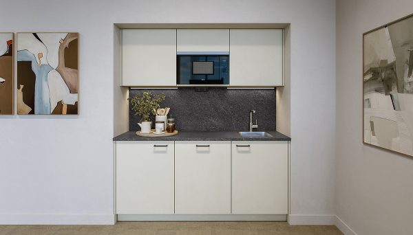 Compact, modern kitchen with light cream-colored cabinet fronts and a clean, symmetrical design. The worktop and backsplash are finished in a dark stone look and are illuminated by integrated under-cabinet lighting. A black microwave is centrally built into the upper cabinets. On the right side, there is a small stainless steel sink with a silver faucet. On the left, white cups, glasses, a green plant, and kitchen accessories in neutral natural tones are arranged decoratively on a tray. The walls are painted in light white and decorated with abstract artwork in beige, brown, and grey tones. The floor is finished with a light, neutral surface.