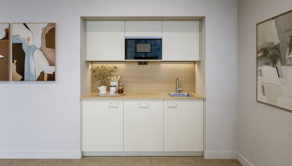 Compact, modern kitchen with light cream-colored cabinet fronts. The worktop and backsplash are finished in a light wood look and are illuminated by under-cabinet lighting. A black microwave is integrated into the upper cabinets. On the right side, there is a small stainless steel sink with a silver faucet. On the left, white cups, transparent glasses, a small green plant, and accessories in neutral, natural tones are arranged on a tray. The walls are painted in light white and decorated with abstract artwork in beige, brown, and grey tones. The floor features a light, neutral surface.