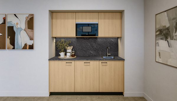 Compact, modern kitchen with cabinet fronts in a light wood finish. The worktop and backsplash are finished in a dark stone look and are evenly illuminated by integrated under‑cabinet lighting. A black microwave is centrally integrated into the upper cabinets. On the right, there is a small stainless steel sink with a silver faucet. On the left, white cups, glasses, a green plant, and kitchen accessories in neutral, natural tones are arranged decoratively on a tray. The walls are painted in light white and decorated with abstract artwork in beige, brown, and grey tones. The floor is finished with a light, neutral surface.