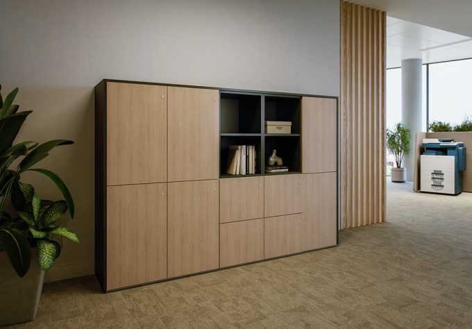 Half‑height locker cabinet in a warm wood finish with a clean, modern design. The cabinet features multiple closed compartments as well as open shelving elements with a dark interior, holding books and a decorative vase. To the left of the cabinet, a large green plant in a planter is placed. The cabinet is integrated into an open office environment with light carpet flooring, light-colored walls, and a partition made of vertical wooden slats in the background.