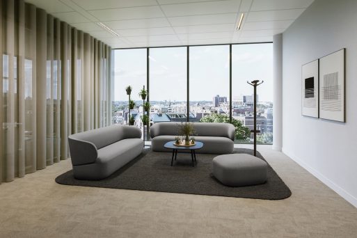 Modern lounge and seating area with floor‑to‑ceiling windows offering views over a cityscape. The space features a seating arrangement consisting of two sofas and a pouf in dark grey tones, combined with a low, round coffee table with a dark tabletop and a slim frame. The seating furniture is arranged on a black rectangular rug, which visually defines the lounge area. On the left, semi‑transparent curtains in a light natural tone hang in front of the windows, while on the right, a simple floor lamp and abstract wall art are visible. Several plants positioned in front of the windows complement the calm, contemporary interior design.