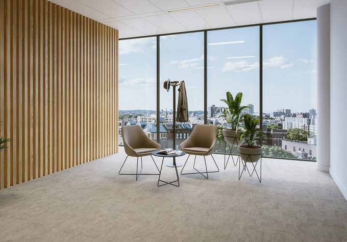 Bright waiting and reception area with two upholstered armchairs in warm brown and sand tones, arranged around a small, round side table with a black metal frame and dark tabletop. The armchairs stand on slim black metal legs. On the left, a floor‑to‑ceiling partition made of light vertical wooden slats creates a warm backdrop. Directly next to the seating area are two plants in grey planters, raised on black metal stands. Behind them, floor‑to‑ceiling window fronts provide ample natural light and offer views of a cityscape. A simple black coat stand completes the modern, welcoming design of the reception area.